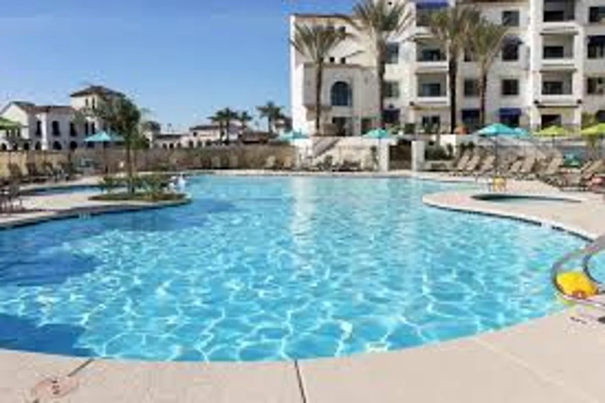 A lively outdoor pool area featuring a sparkling swimming pool, colorful umbrellas, lounge chairs, and palm trees, buzzing with activity as residents swim and socialize. The overall vibe is cheerful and community-focused.