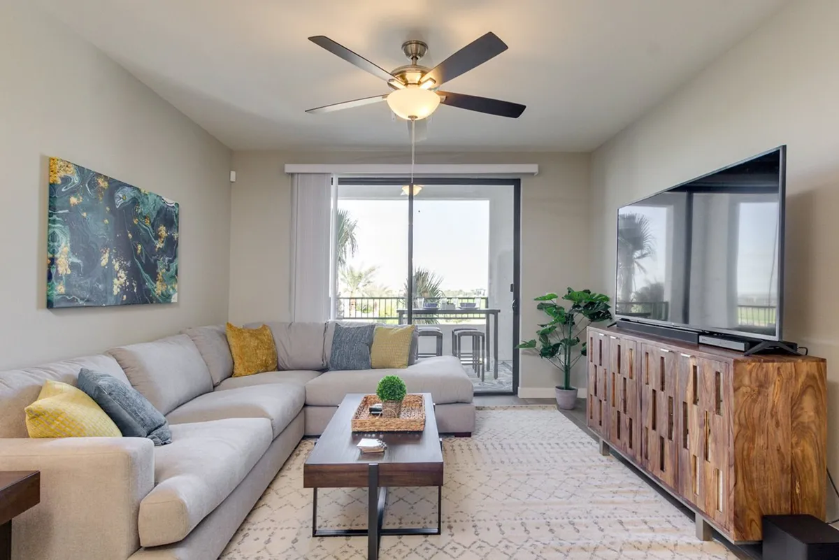 The image showcases a comfortable and stylish open-concept living space, with cozy sofas, a coffee table, modern artwork, and a television. The setting suggests a relaxing environment for socializing, watching TV, or enjoying a sunset from the balcony, reinforcing leisure and upscale comfort.