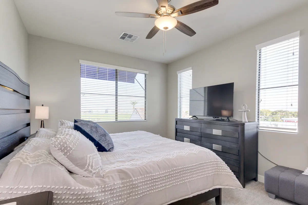 A cozy bedroom scene with a king bed adorned with decorative pillows, a television atop a dresser, and abundant natural light streaming in from the windows. The space feels inviting—ideal for unwinding or enjoying a quiet movie night.