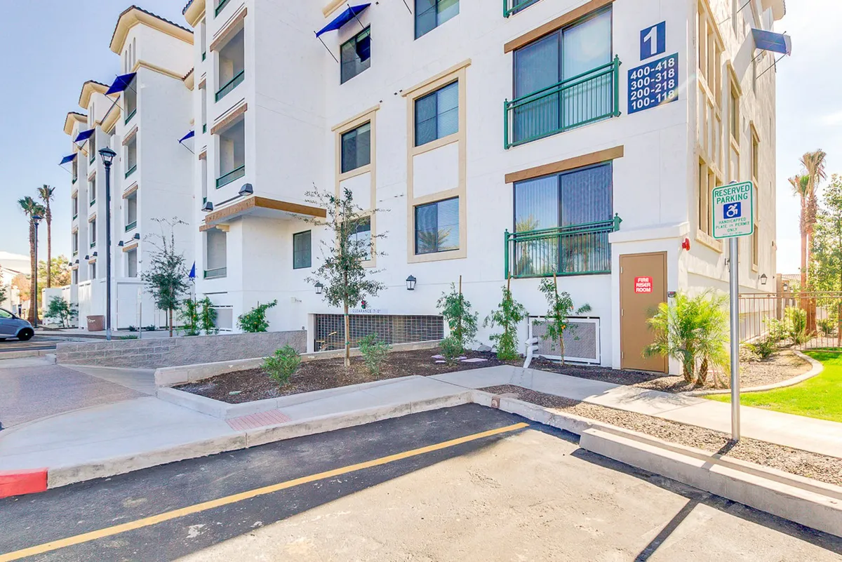 A welcoming exterior perspective showcasing the building’s facade, entrance area, lush green space, and nearby parking. The setting is ideal for residents to gather, relax, or enjoy morning coffee in the open air.