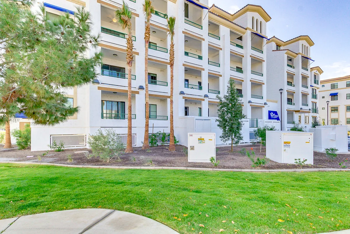 A wide exterior view showcasing the modern condo building surrounded by palm trees and a lush green lawn. The setting is serene and inviting, with thoughtful landscaping and a sense of community life. The image suggests opportunities for outdoor relaxation or a pleasant stroll, echoing the vibrancy of the local area.