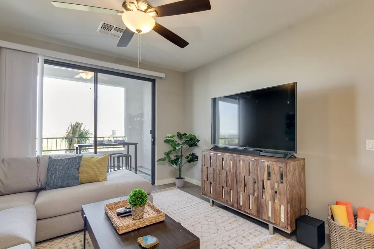A comfortable living room scene with a plush sofa facing a large wall-mounted 4K TV, a coffee table, greenery, and a balcony view letting in sunset light. The setup is perfect for relaxing, watching shows, or entertaining friends, with a cozy yet contemporary ambiance.