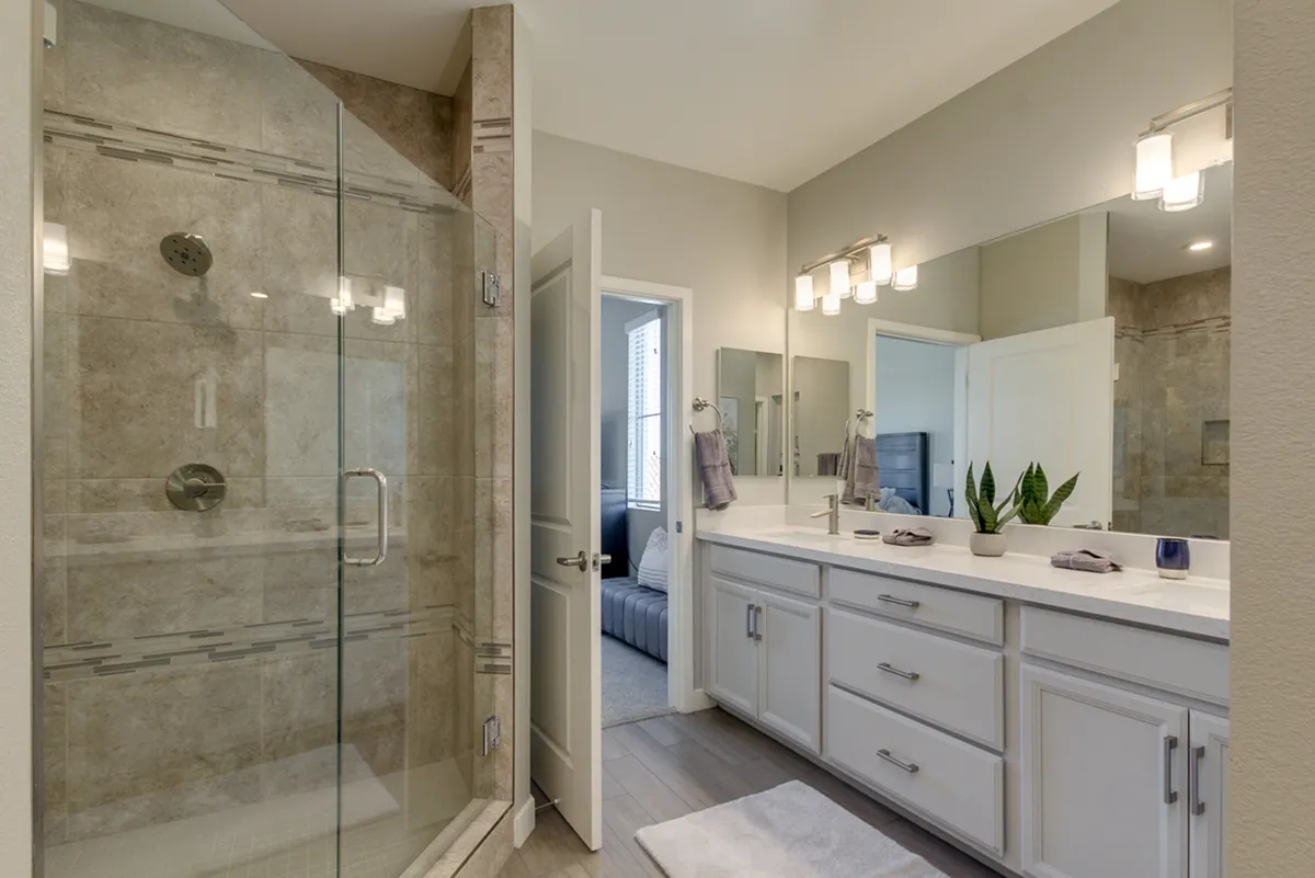 A spa-inspired bathroom with a sleek glass walk-in shower, double quartz sink vanity, mirrors, and a touch of greenery. The clean lines and tranquil design evoke a sense of modern luxury and relaxation.