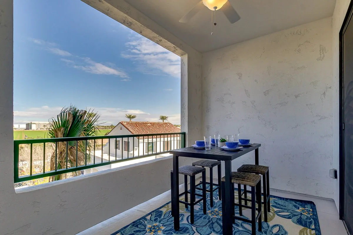 A stylish exterior balcony setup with a dining table, decorative rug, and sweeping views of palm trees and distant buildings. The space is arranged for al fresco dining or morning coffee, capturing a tranquil, resort-style outdoor experience.