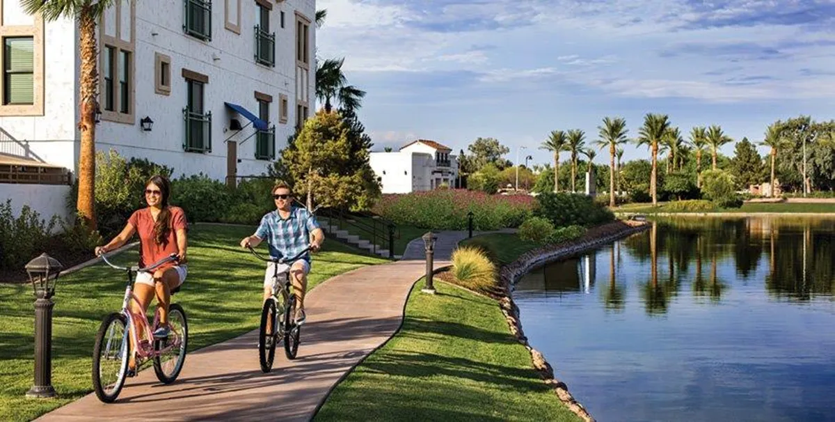 A vibrant exterior scene featuring bicyclists on a waterfront path lined with palm trees and green space. The area is alive with people enjoying nature, suggesting an active, community-oriented atmosphere.
