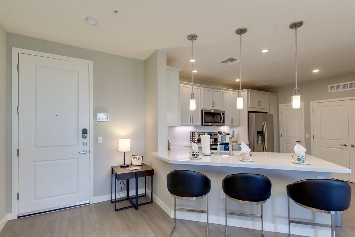 A bright kitchen scene featuring a quartz-topped island with bar stools, stylish lighting, and sightlines to the entrance. The space is designed for both culinary activity and social interaction, making it ideal for coffee at the breakfast bar or hosting guests.