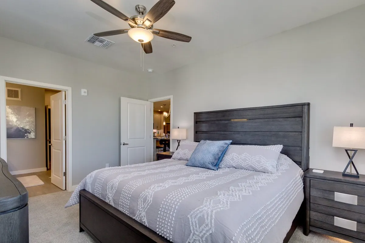 This image features a serene bedroom with a king-size bed, plush bedding, a nightstand with a lamp, and a ceiling fan. The neutral color palette and soft lighting evoke a calm, tranquil mood perfect for restful sleep.