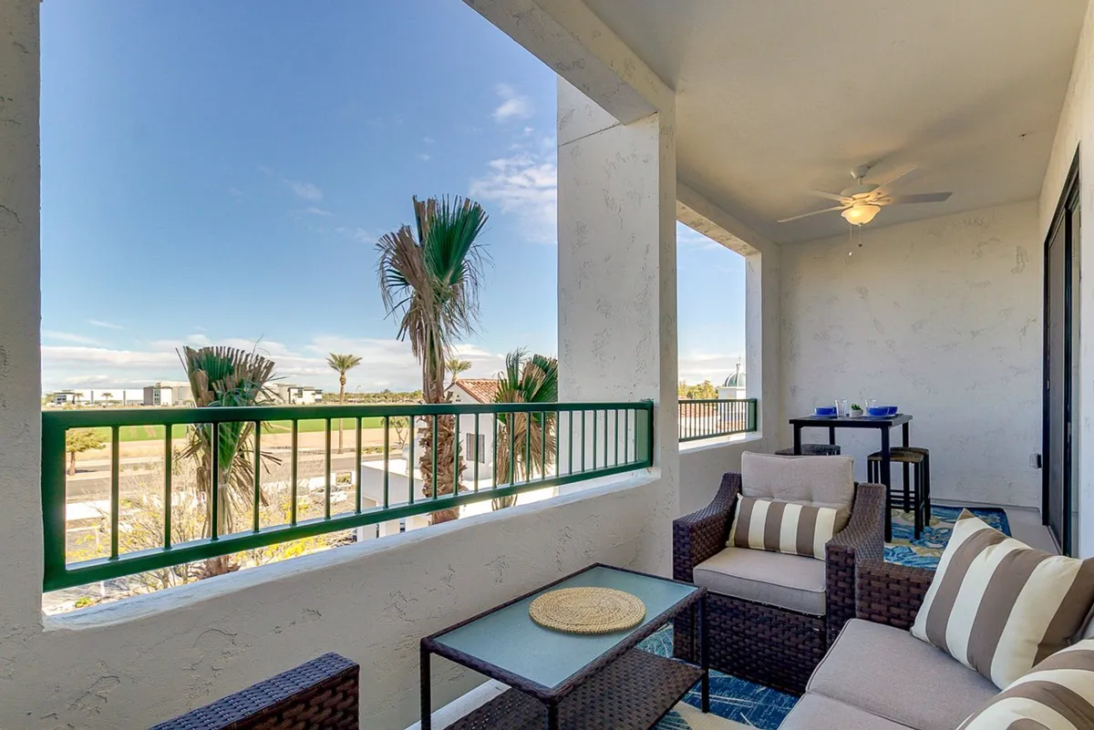 A spacious, comfortable outdoor seating area with cushioned chairs, an outdoor table, and green railing. Palm trees provide a lush, resort-like backdrop, and the setup is ideal for relaxing, entertaining guests, or enjoying morning coffee while overlooking the landscape.