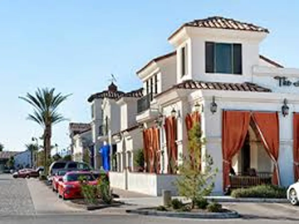 A lively exterior scene showing a building with bright orange curtains and palm trees, where residents can be seen strolling along the street, sitting on benches, and socializing with neighbors. The presence of parked cars adds to the sense of an accessible, walkable community.